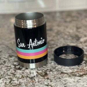 K&ouml;nnte beinhalten: Ein schwarzer, isolierter Getr&auml;nkedosenk&uuml;hler aus Edelstahl mit einer wei&szlig;en Grafik, auf der "San Antonio" steht, und bunten Streifen. Der K&uuml;hler ist offen und der Deckel liegt daneben.