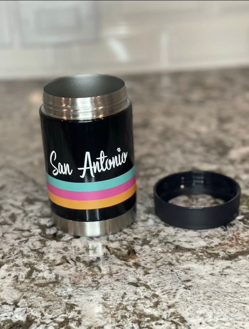 K&ouml;nnte beinhalten: Ein schwarzer, isolierter Getr&auml;nkedosenk&uuml;hler aus Edelstahl mit einer wei&szlig;en Grafik, auf der "San Antonio" steht, und bunten Streifen. Der K&uuml;hler ist offen und der Deckel liegt daneben.
