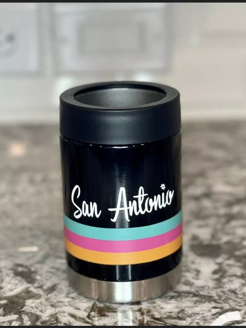 K&ouml;nnte beinhalten: Schwarzer und silberner Getr&auml;nkedosenk&uuml;hler mit einer wei&szlig;en Grafik, die "San Antonio" mit einem Stern &uuml;ber dem "o" sagt. Die Grafik ist &uuml;ber drei horizontale Streifen in Orange, Pink und T&uuml;rkis gelegt.