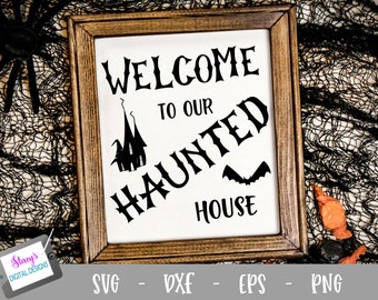 May include: A rectangular wooden frame containing a white sign with black text that reads WELCOME TO OUR HAUNTED HOUSE. The design includes a black silhouette of a tall, pointed house and a flying bat. The frame is set against a black mesh fabric.