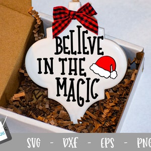 May include: White ceramic ornament with a red and black checked bow, the text "Believe in the Magic" and a red Santa hat. The ornament is in a box with brown packing paper.
