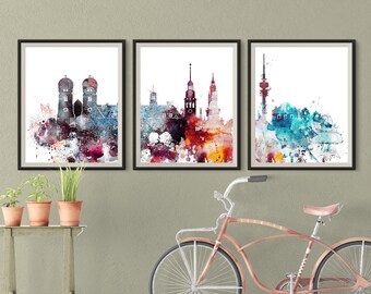 Puede incluir: Tres acuarelas enmarcadas de paisajes urbanos. La primera pintura es un paisaje urbano rojo y azul con dos edificios altos. La segunda pintura es un paisaje urbano rojo, naranja y amarillo con un edificio alto de iglesia. La tercera pintura es un paisaje urbano azul y verde con una torre alta.