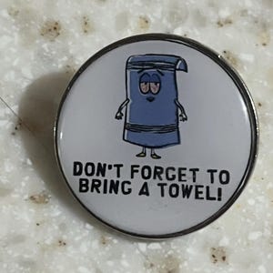 May include: A white round pin with a silver border. The pin features a cartoon illustration of a blue towel with a face and the text "Don't forget to bring a towel!"