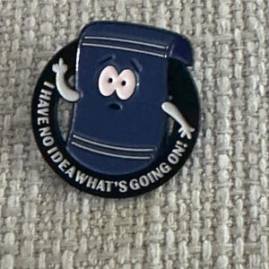May include: A blue enamel pin featuring a cartoon towel character with wide eyes and raised arms. The pin is surrounded by a black circle with the text "I HAVE NO IDEA WHAT'S GOING ON!" in white lettering. The towel character has a surprised expression.