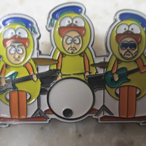 May include: A yellow, blue, and white enamel pin featuring three cartoon characters dressed as chickens playing musical instruments. The character on the left is playing a blue electric guitar, the character in the middle is playing a drum set, and the character on the right is playing a green electric guitar.