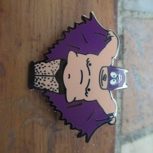 May include: A purple and black enamel pin featuring a cartoon character with a pink body, black eyes, and a purple mask. The character is wearing a purple cape and has a black and white patterned belt.