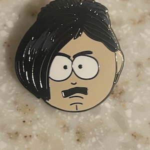 May include: A black and white enamel pin featuring a cartoon character with black hair, a mustache, and a grumpy expression.