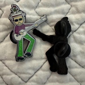 May include: A metal enamel pin featuring a figure wearing a white hat with a small green creature on top, sunglasses, a purple striped shirt, and glittery green pants while playing a white guitar, shown alongside its black metal backing piece.
