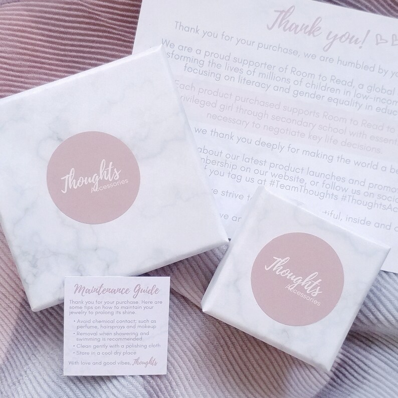 May include: White marble-patterned boxes and a small card with the text "Thoughts Accessories" in a soft pink circle. A "Thank you" card and a "Maintenance Guide" card are also visible. The items are on a pink and white striped surface.