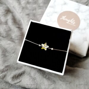 May include: A silver bracelet with a star-shaped charm, presented in a black box. The star charm is yellow and clear, with small beads on the bracelet. A white and marble-patterned box is in the background.