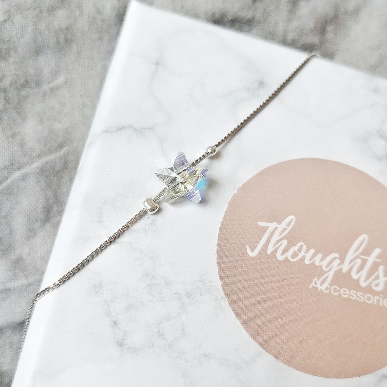 May include: A delicate silver bracelet featuring a sparkling, multi-faceted star pendant. The star has iridescent qualities, reflecting various colors. The bracelet is set against a marble-patterned background, with the text "Thoughts Accessories" visible.