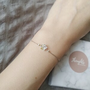 May include: A delicate silver bracelet featuring a floral charm with clear and yellow crystals. The bracelet is worn on a wrist, with a box in the background that has the word "Thoughts" on it.