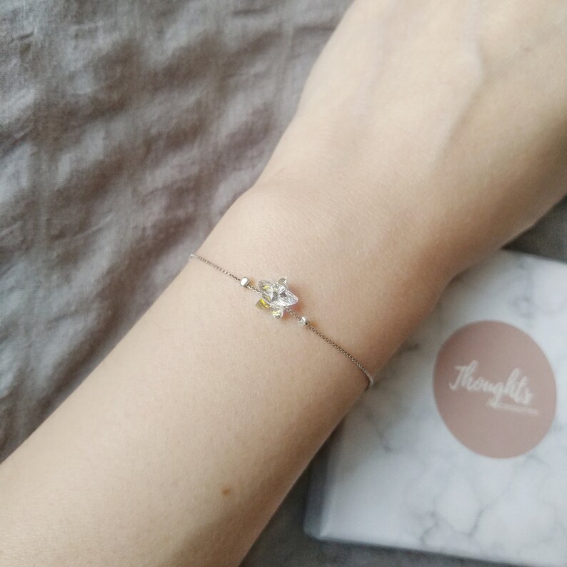 May include: A delicate silver bracelet featuring a floral charm with clear and yellow crystals. The bracelet is worn on a wrist, with a box in the background that has the word "Thoughts" on it.