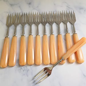 May include: A set of ten vintage forks with stainless steel tines and a light orange, marbled handle. One fork is placed separately. The forks are arranged on a white and gray marble surface.