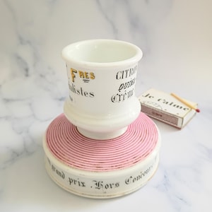 K&ouml;nnte beinhalten: Ein wei&szlig;er Keramik-Streichholzhalter mit rosa Sockel und gold-schwarzem Text. Der Text enth&auml;lt die W&ouml;rter "FRES", "CITRON" und "grand prix Hors Concours". Eine Streichholzschachtel mit dem Text "Je t'aime" liegt daneben.