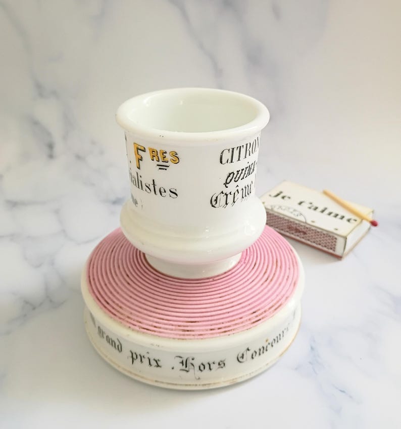 K&ouml;nnte beinhalten: Ein wei&szlig;er Keramik-Streichholzhalter mit rosa Sockel und gold-schwarzem Text. Der Text enth&auml;lt die W&ouml;rter "FRES", "CITRON" und "grand prix Hors Concours". Eine Streichholzschachtel mit dem Text "Je t'aime" liegt daneben.