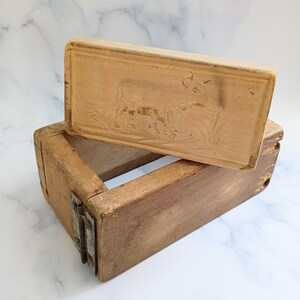 May include: A vintage wooden butter mold with a rectangular shape. The lid features a carved deer design. The mold is open, revealing the interior. The wood has a warm, natural tone, and the metal hinges are visible.