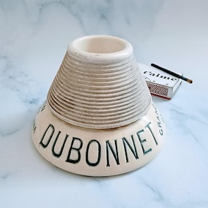 May include: A cream-colored ceramic Dubonnet advertising ashtray with a wide, sloped base featuring the black text DUBONNET and a raised, ribbed conical center section. A small matchbox with the text t'aime and a single matchstick are visible.