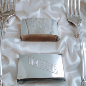 May include: Two silver napkin rings with the words "MADAME" and "MONSIEUR" engraved on them. The rings are on a white satin surface.