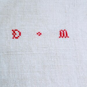 May include: A piece of off-white, woven fabric featuring a cross-stitched monogram in red thread. The embroidery displays the capital letter D, a small decorative diamond-shaped cross, and the lowercase letter m, all centered on the textile.