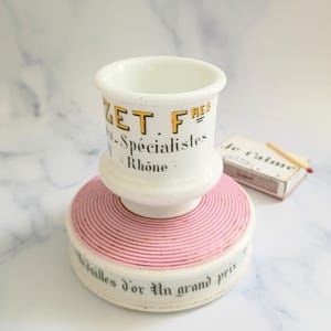 K&ouml;nnte beinhalten: Ein wei&szlig;er Keramik-Streichholzhalter mit rosa Sockel und passender Streichholzschachtel. Der Streichholzhalter tr&auml;gt den Text "ZET FRES - Sp&eacute;cialistes Rh&ocirc;ne" in Gelb und Schwarz. Der Sockel tr&auml;gt den Text "M&eacute;dailles d'or Un grand prix".