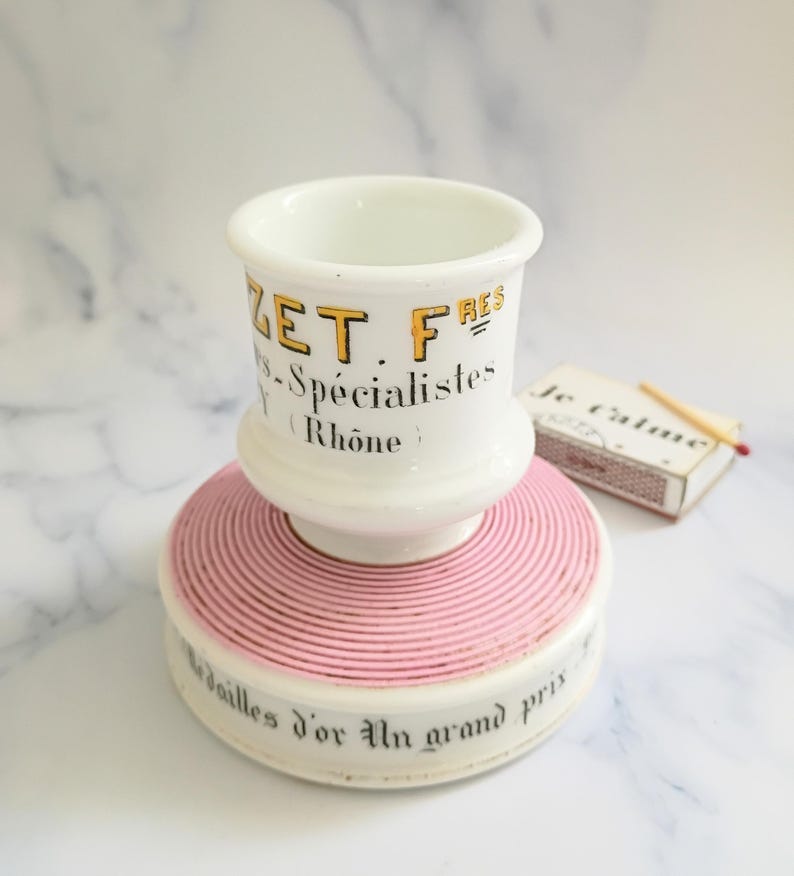 K&ouml;nnte beinhalten: Ein wei&szlig;er Keramik-Streichholzhalter mit rosa Sockel und passender Streichholzschachtel. Der Streichholzhalter tr&auml;gt den Text "ZET FRES - Sp&eacute;cialistes Rh&ocirc;ne" in Gelb und Schwarz. Der Sockel tr&auml;gt den Text "M&eacute;dailles d'or Un grand prix".