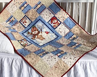 May include: A patchwork quilt featuring a central snowman illustration surrounded by blue and beige patterned squares with a red border, displayed on a white crib alongside a plush gnome with a tall red hat, white beard, and gray body.