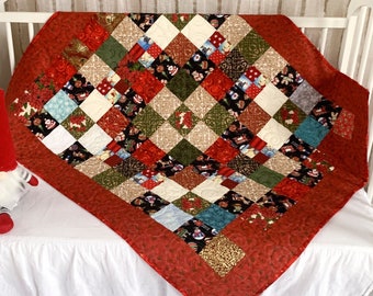 May include: A patchwork quilt featuring a variety of holiday-themed fabric squares in red, green, gold, and white, arranged in a diamond pattern. The quilt has a solid red border with a swirling quilted texture and is displayed on a white crib.