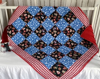 May include: A square patchwork quilt featuring a diamond-patterned design with alternating blocks of blue fabric with white snowflakes and black fabric with colorful Christmas-themed icons, finished with a red and white checkered border.