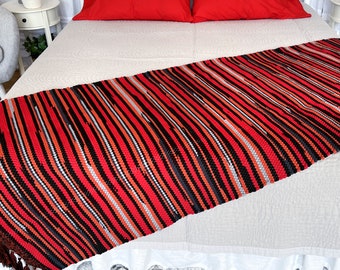 May include: A rectangular bed runner featuring a woven striped pattern in shades of red, black, and grey. The textile has a textured surface with vertical lines and dark brown fringe tassels attached to the short ends of the fabric piece.