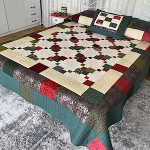 Op de afbeelding: Een patchwork sprei met een raster van crèmekleurige, donkerrode, bosgroene en bruine stoffen vierkanten met bloem- en krulpatronen. De sprei ligt over een bed met een bijpassend vierkant kussen met een centraal ontwerp van negen vlakken.