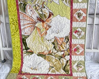 May include: A quilted baby blanket featuring a central illustration of a fairy with wings sitting among large white flowers and green foliage. The border includes a patchwork design of small floral squares in shades of orange, green, and red.
