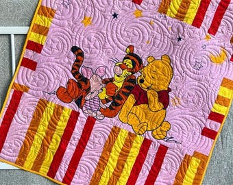 May include: A square quilt featuring a central light pink panel with embroidered Winnie the Pooh, Tigger, and Piglet characters, surrounded by a border of alternating red, orange, and yellow vertical stripes with intricate swirl stitching.