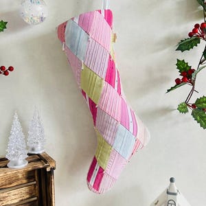 Könnte beinhalten: Ein gesteppter Weihnachtsstrumpf mit einem Patchwork-Design aus diagonalen Streifen in Rosa-, Hellblau- und Gelbtönen. Der Strumpf besteht aus Stoffstreifen mit sichtbaren Nähten und hängt an einer Schlaufe aus weißem Band.
