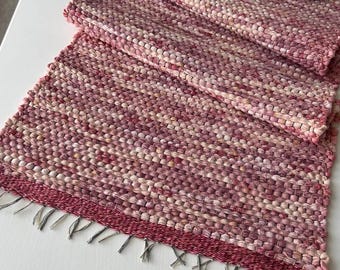 May include: A rectangular woven table runner featuring a variegated pattern of pink, magenta, and cream-colored yarns. The runner has a thick, textured weave and short, frayed fringe edges. A dark brown ceramic cup and saucer sit on the runner.