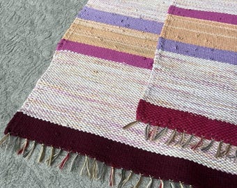 May include: Two rectangular woven rugs featuring horizontal stripes in shades of deep burgundy, light purple, soft orange, and cream. The rugs have a textured, multi-colored weave and are finished with short, knotted fringe along the bottom edges.