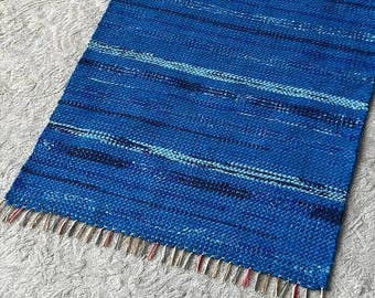 May include: A rectangular woven rug featuring a vibrant blue color palette with horizontal stripes in varying shades of navy, royal blue, and light blue. The rug has a textured, braided weave pattern and includes short, frayed fringe edges.