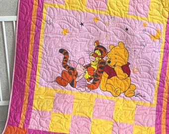 May include: A quilted blanket featuring a central panel with embroidered illustrations of Winnie the Pooh, Tigger, and Piglet under a night sky with stars and a crescent moon. The border consists of a pink and yellow checkered pattern with magenta trim.