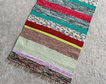 May include: A rectangular woven rag rug featuring a vibrant horizontal striped pattern in shades of mint green, bright pink, teal, neon yellow, brown, and purple. The textile has a textured, multi-colored appearance with short fringe at both ends.