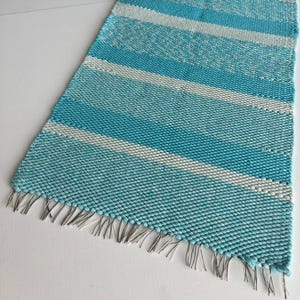 May include: A rectangular woven table runner featuring horizontal stripes in alternating shades of turquoise and white. The fabric has a textured, chunky knit appearance and includes short, thin fringe detailing along the two shorter ends.