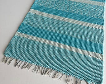 May include: A rectangular woven table runner featuring horizontal stripes in alternating shades of turquoise and white. The fabric has a textured, chunky knit appearance and includes short, thin fringe detailing along the two shorter ends.