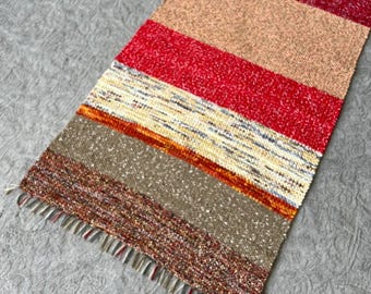 May include: A rectangular woven runner rug featuring horizontal stripes in various colors including light green, yellow, deep red, tan, bright red, cream with orange flecks, brown, and dark brown. The rug has a textured finish and fringe.