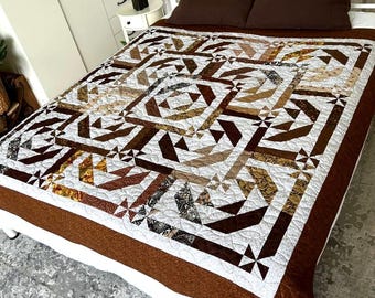 May include: A rectangular quilt featuring a geometric pattern in shades of brown, tan, cream, and white. The design includes intricate pieced blocks with star-like motifs and a solid dark brown border, finished with detailed overall quilting stitches.
