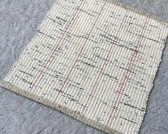 May include: A rectangular woven rug featuring a cream-colored base with a textured surface. The design includes vertical stripes in thin red and dark gray lines, with a solid tan border along the top and bottom edges of the fabric piece.