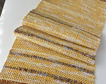 May include: A rectangular woven table runner featuring a textured pattern of yellow, white, and muted red fabric strips. The runner has a thick, dark red border at the ends and includes decorative gray fringe tassels along the short edges.