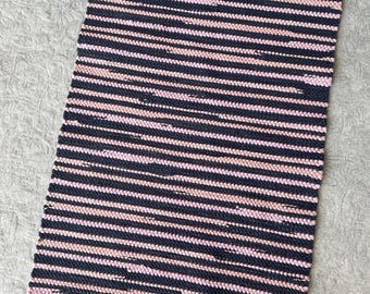 May include: A rectangular woven rug featuring a repeating pattern of horizontal stripes in dark navy blue and soft pastel pink. The rug is finished with short, frayed fringe detailing along the top and bottom edges, creating a textured appearance.
