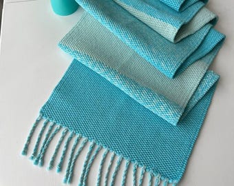 May include: A rectangular table runner featuring a textured weave in shades of bright turquoise and pale mint green. The fabric displays a gradient color transition and is finished with braided fringe tassels along the short edges.