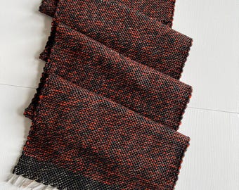 May include: A rectangular woven table runner featuring a dense, textured pattern of intertwined black and deep red threads. The fabric displays a consistent, tight weave throughout its length, finished with short, light-colored fringe at the ends.
