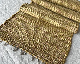 May include: A rectangular table runner featuring a woven texture in shades of mustard yellow, gold, and hints of brown. The runner has a thick, chunky weave pattern and is finished with short, thin, light-colored fringe tassels along the bottom edge.