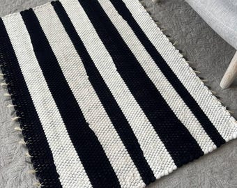 May include: A rectangular woven rug featuring a bold pattern of thick, alternating black and white vertical stripes. The rug has a textured, chunky knit appearance with visible fringe detailing along the side edges, creating a high-contrast design.